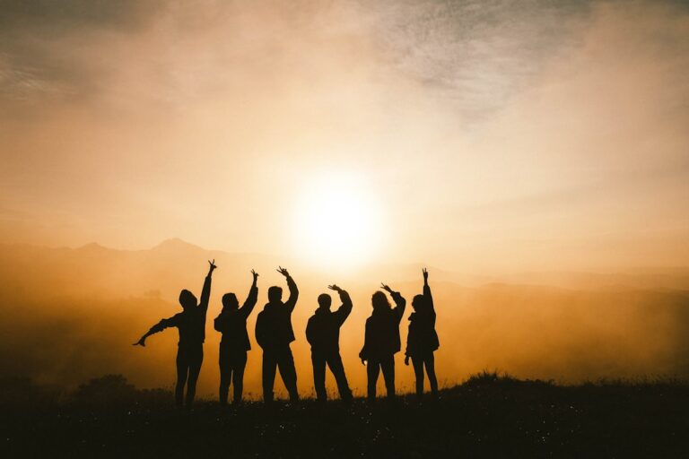 Silhouette of a group of six people raising their hands together at sunrise, symbolizing peer support and community in recovery