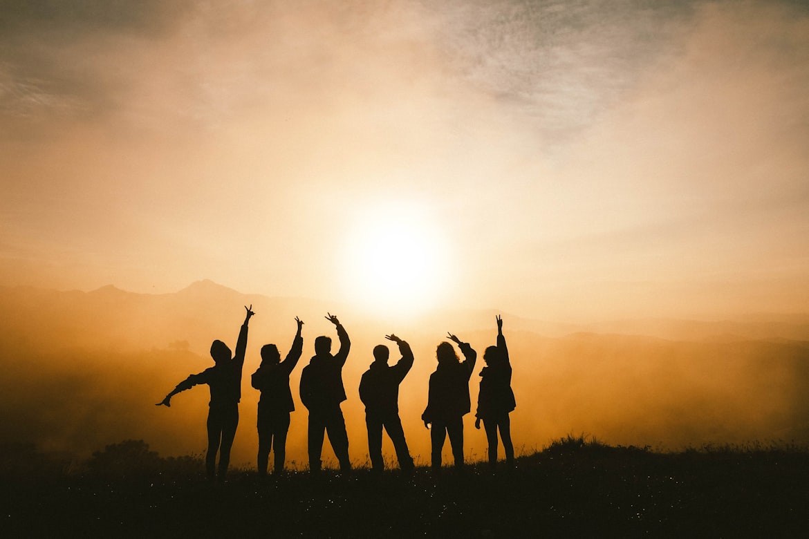 Silhouette of a group of six people raising their hands together at sunrise, symbolizing peer support and community in recovery